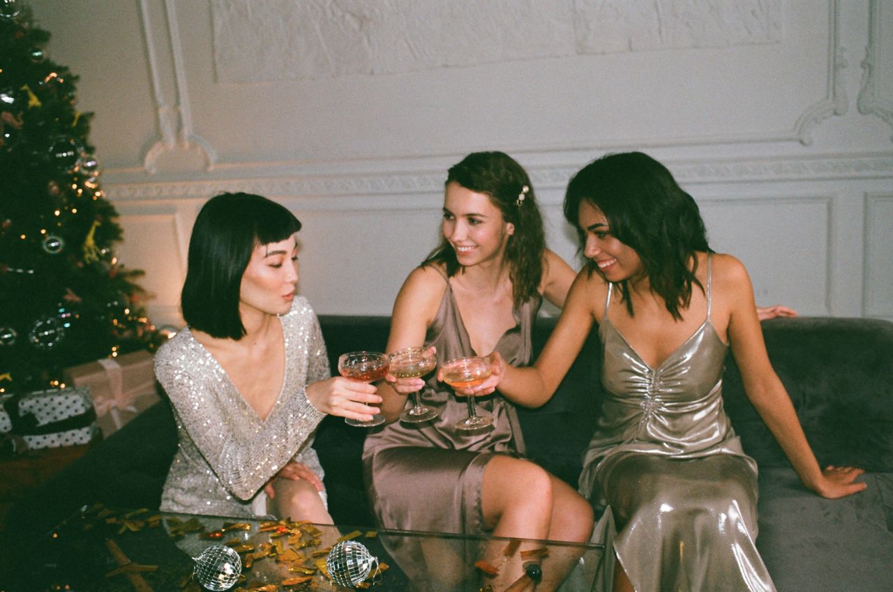 creative-02 Three friends in glamorous dresses sharing drinks at a festive indoor gathering.